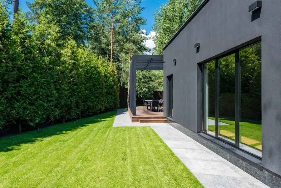 A grey home beside a lawn, with a timber pergola and tall hedges